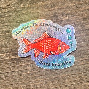 Puede incluir: Una pegatina holográfica con un pez dorado rojo y el texto "Anxious Goldfish, says... Just breathe." La pegatina tiene un acabado brillante e iridiscente y está sobre un fondo de madera.