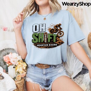 Könnte beinhalten: Hellblaues T-Shirt mit dem Text "OH SHIFT" in Weiß und Grün sowie einer Mountainbike-Grafik. Die Grafik zeigt einen Radfahrer auf einem Mountainbike. Die Worte "MOUNTAIN BIKING" stehen unter der Grafik.