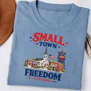 May include: A dusty blue t-shirt featuring a graphic print of a small town scene with a church, a water tower, and shops. The text reads "SMALL TOWN FREEDOM" in red and blue, with "1776-2026" printed below, accented by small red stars.