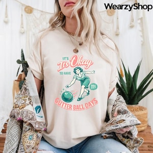 May include: Cream-colored t-shirt with a retro bowling graphic. The design features a woman bowling, a teal bowling ball, and the text "It's Okay to Have Gutter Ball Days" in pink and teal. The shirt has short sleeves.