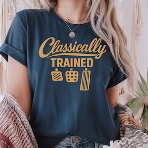 Könnte beinhalten: Marineblaues T-Shirt mit dem Text "Classically Trained" in goldener Schrift über gelben Pedal-Grafiken. Das Shirt hat hochgekrempelte Ärmel und ist aus einem weichen Material.