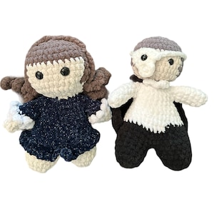 May include: Two handmade crochet dolls. One doll has brown hair and a navy blue dress. The other doll is dressed in black and white, with a grey mask. Both dolls have black button eyes and are made of soft yarn.