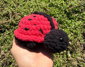 May include: A crocheted ladybug toy with a red body, black spots, and black head and legs. The ladybug is held in a person's hand.