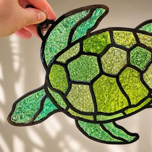 May include: A stained glass sea turtle suncatcher featuring a mosaic of textured green and lime glass pieces held together by dark metal solder lines. The turtle shape includes a detailed shell pattern and four flippers, with a small metal loop.