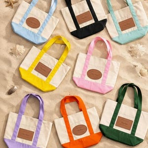 May include: A collection of small canvas tote bags in various colors, including blue, black, yellow, pink, purple, orange, and green. Each bag has a cream-colored body, colored accents, and a brown leather patch. The bags are arranged on a sandy surface.