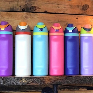 May include: A collection of colorful insulated water bottles displayed on a wooden shelf. The bottles come in various colors, including pink, red, blue, and white, with matching lids. Each bottle is approximately 10 inches tall.