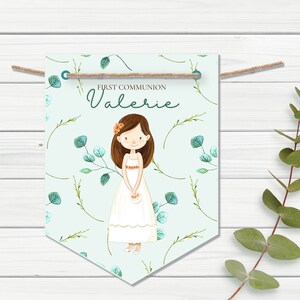 May include: A light blue banner with a white border and a floral pattern. The banner has the text "FIRST COMMUNION Valerie" and a cartoon illustration of a girl in a white dress holding a basket of flowers.