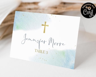 May include: A white place card with watercolor blue and green accents and gold flecks. The card features a gold cross and the name "Jennifer Moore" in elegant script, with "TABLE 3" below.