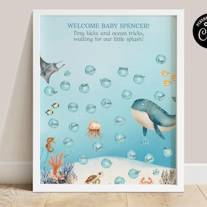 Op de afbeelding: Een ingelijste baby shower print met een oceaan thema. Het kunstwerk toont een walvis, een manta rog, een zeepaardje en andere zeedieren. Bubbels met namen zweven in het blauwe water, met de tekst "WELCOME BABY SPENCER!" bovenaan.