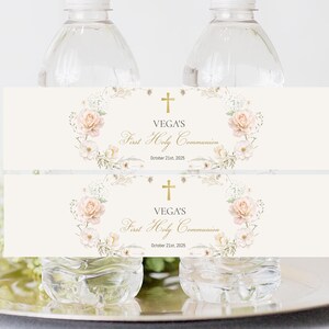 Puede incluir: Dos botellas de agua con etiquetas para la Primera Comunión. Las etiquetas son de color crema con un diseño de corona floral, una cruz dorada y el texto "VEGA'S First Holy Communion October 21st, 2025."