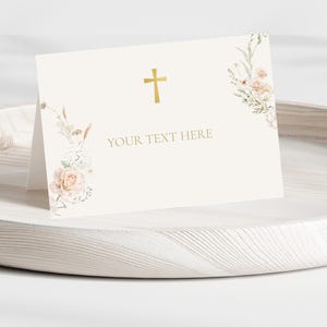 May include: A folded cream card featuring a gold cross and the text "YOUR TEXT HERE". It's adorned with floral arrangements in soft pink and cream hues. The card is presented on a light wooden tray.