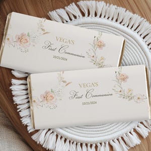 Puede incluir: Dos envoltorios de barra de chocolate blanco con diseños florales y letras doradas. El texto dice "VEGA'S First Communion 10/21/2024".