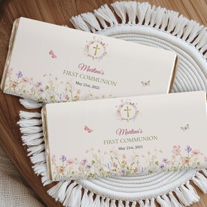 Puede incluir: Dos envoltorios de barra de chocolate blanco con un diseño floral y una cruz dorada. El texto en los envoltorios dice "Martina's First Communion May 21st, 2025".