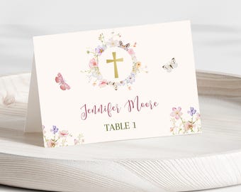 May include: A white place card with a floral wreath design featuring pink, purple, and yellow flowers. A gold cross is in the center of the wreath. The text "Jennifer Moore" and "Table 1" is printed in pink and green. Butterflies are also included in the design.