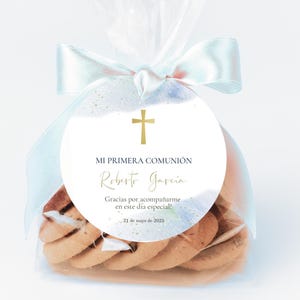 Puede incluir: Una pegatina circular de estilo acuarela blanca y azul con una cruz dorada y el texto "MI PRIMERA COMUNIÓN Roberto Garcia Gracias por acompañarme en este día especial! 21 de mayo de 2025". La pegatina está pegada a una bolsa de celofán transparente llena de galletas.
