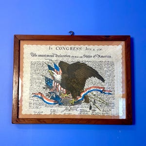 May include: Framed print of the Declaration of Independence, featuring an eagle with outstretched wings, American flags, and red, white, and blue ribbons. The print is in a dark wood frame and hangs against a blue wall.