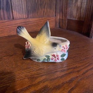 May include: A yellow bird figurine with black wings and a brown head, perched on a flower pot with pink flowers and green leaves.