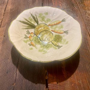 May include: A white ceramic serving bowl with a scalloped edge, decorated with hand-painted vegetables. The design includes green zucchini slices, carrots, and green leafy accents. The bowl sits on a wooden surface.