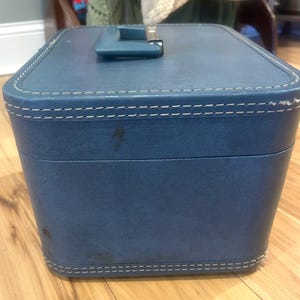 May include: A rectangular blue vanity case with a top handle and white contrast stitching along the edges. The exterior features a smooth, textured finish with visible signs of wear, including small dark spots on the front and side panels.