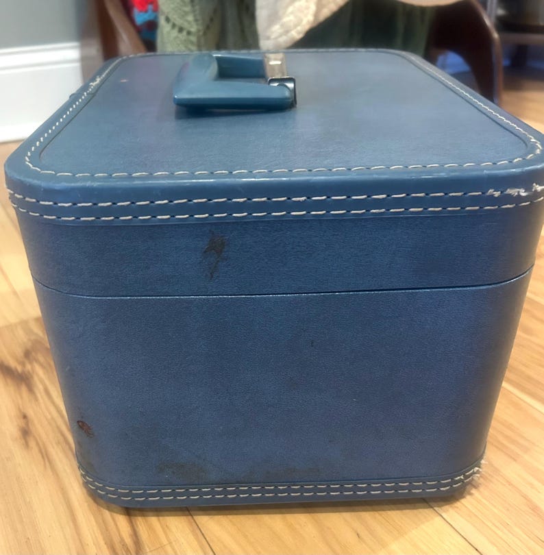 May include: A rectangular blue vanity case with a top handle and white contrast stitching along the edges. The exterior features a smooth, textured finish with visible signs of wear, including small dark spots on the front and side panels.