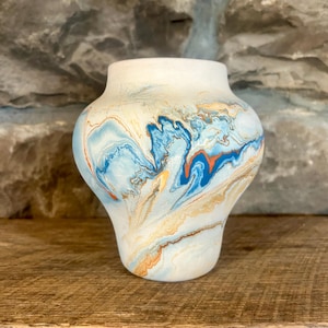 May include: A ceramic vase with a white rim and a body decorated with swirling blue, orange, and tan patterns. The vase has a rounded shape and sits on a wooden surface against a stone wall.