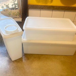 May include: A set of white plastic food storage containers with lids. The containers are rectangular and stackable, including a tall, narrow container and two wider, shorter ones. The lids are white and fit securely on the containers.