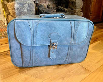 May include: A vintage blue suitcase with a silver clasp featuring a peace symbol. The suitcase has a handle and is made of a textured material. Vertical stitching details the front and sides. The suitcase is on a wooden floor.