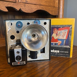 May include: A vintage Brownie Hawkeye camera with a flash attachment in a black box. The box includes a flash bulb and a flash unit. The camera is black with silver accents and says "Brownie Hawkeye Flash Outfit" on the front. The box has a yellow lid with the words "Hawkeye Flash Outfit" and a picture of a girl painting.