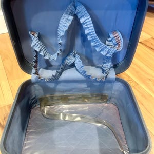 May include: A vintage blue hard-shell train case with a hinged lid and metal latches. The interior features a light blue fabric lining, a gathered elastic pocket along the upper lid, and a quilted base with a clear plastic strap across the bottom.