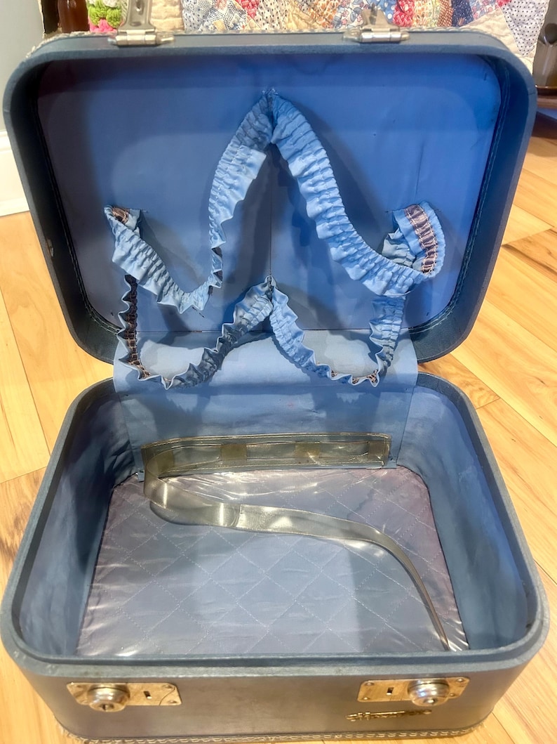 May include: A vintage blue hard-shell train case with a hinged lid and metal latches. The interior features a light blue fabric lining, a gathered elastic pocket along the upper lid, and a quilted base with a clear plastic strap across the bottom.