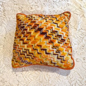 May include: A square throw pillow featuring a textured needlepoint design with a repeating chevron pattern in shades of orange, yellow, cream, and dark brown. The pillow is finished with a solid orange fabric piping along all four edges.