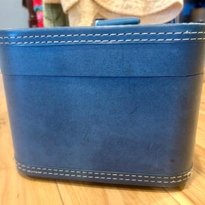 May include: A rectangular blue vanity case with rounded corners and a smooth finish. The lid and base feature prominent white contrast stitching along the edges. A small blue handle is attached to the top center of the lid for carrying.