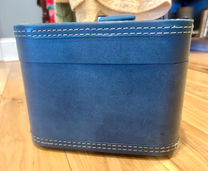May include: A rectangular blue vanity case with rounded corners and a smooth finish. The lid and base feature prominent white contrast stitching along the edges. A small blue handle is attached to the top center of the lid for carrying.
