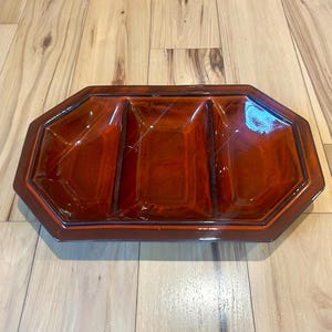May include: A rectangular, divided serving dish with a glossy, reddish-brown finish. The dish has three compartments and a geometric, octagonal shape. The edges are slightly raised, and the dish sits on a wooden surface.