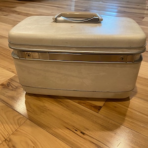 May include: Cream-colored vintage vanity case with a textured finish. The rectangular case features a silver-toned metal trim and handle. The case has two latches and a keyhole on the front. The handle is a light beige color.