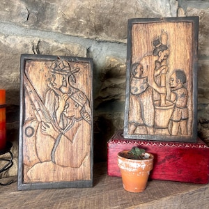 May include: Two wooden relief carvings depicting figures in traditional clothing. One shows a man with a rifle, the other shows a woman and children. The carvings are framed in dark wood and rest on a wooden shelf.