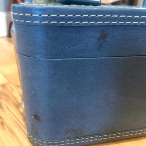 May include: A blue hard-sided vanity case featuring a top handle and white contrast stitching along the edges. The exterior surface displays visible signs of wear, including small dark spots and scuffs on the front panel of the rectangular container.