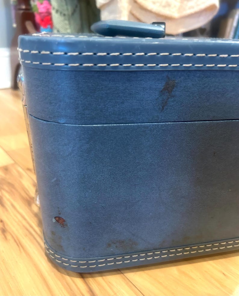 May include: A blue hard-sided vanity case featuring a top handle and white contrast stitching along the edges. The exterior surface displays visible signs of wear, including small dark spots and scuffs on the front panel of the rectangular container.