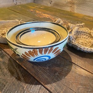 May include: Three nested ceramic bowls with a white background and colorful floral designs. The bowls are on a wooden surface with two woven baskets.