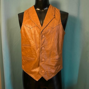 May include: A brown leather waistcoat with a V-neck and four buttons. The waistcoat has two front pockets and decorative stitching. It is displayed on a black mannequin. The colour is a warm tan.
