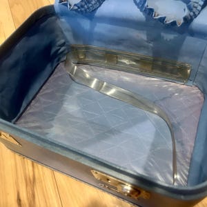 May include: An open, rectangular light blue vanity case with a quilted interior lining. The case features a metallic handle attached to the inner lid, gold-toned metal latches on the exterior sides, and a smooth, light blue fabric interior.