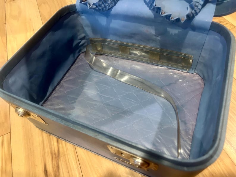 May include: An open, rectangular light blue vanity case with a quilted interior lining. The case features a metallic handle attached to the inner lid, gold-toned metal latches on the exterior sides, and a smooth, light blue fabric interior.