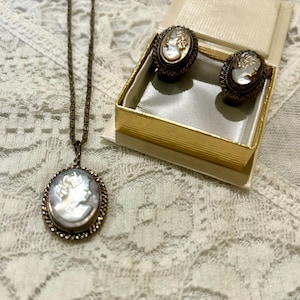 May include: A vintage jewelry set featuring a cameo pendant necklace and matching earrings. The oval pendant and earrings showcase a white cameo set in a bronze-toned metal frame. The earrings are displayed in a gold box on a lace surface.