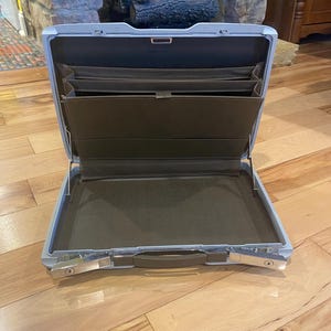 May include: Open, light blue briefcase with a silver clasp and handle. The interior features brown compartments and a large open space. The briefcase is designed for carrying documents and other business essentials.