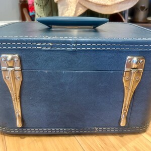 May include: A rectangular blue train case with a matching blue handle on top. The case features white contrast stitching along the edges and two vertical silver-toned metal latches on the front. The exterior has a smooth, solid-colored finish.