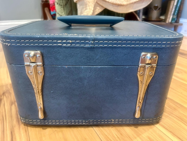 May include: A rectangular blue train case with a matching blue handle on top. The case features white contrast stitching along the edges and two vertical silver-toned metal latches on the front. The exterior has a smooth, solid-colored finish.