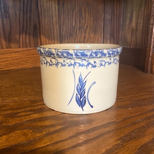 May include: A white ceramic crock with a blue and white floral design. The crock has a blue wheat design on the front.