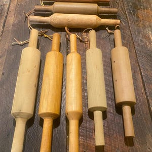 May include: An assortment of wooden rolling pins in shades of light brown and tan, displayed on a rustic wooden surface. Some pins have leather loops. The rolling pins vary in size and shape, showing natural wood grain.