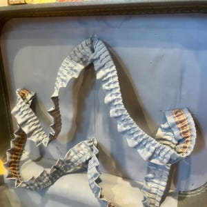 May include: The interior of a vintage suitcase featuring a light blue fabric lining. A decorative, ruffled light blue fabric trim with a thin brown striped accent runs along the inner lid, creating a gathered, wavy border against the smooth surface.