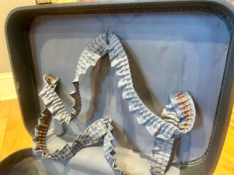 May include: The interior of a vintage suitcase featuring a light blue fabric lining. A decorative, ruffled light blue fabric trim with a thin brown striped accent runs along the inner lid, creating a gathered, wavy border against the smooth surface.